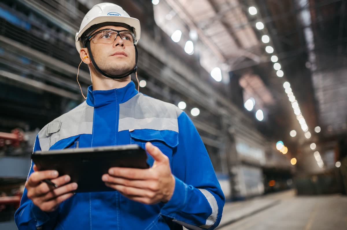An inspector in a hard hat and high-visibility jacket holding a tablet in a warehouse