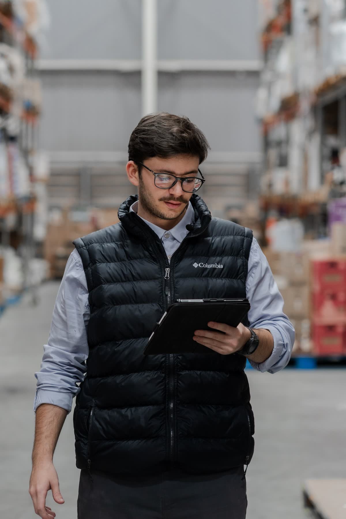 A man in glasses and a quilted vest checking a tablet in a warehouse aisle